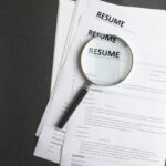 Recruiters-workplace-pile-of-resume-examples-hand-holding-magnifier-Concept-of-analyzing-information-about-new-employees